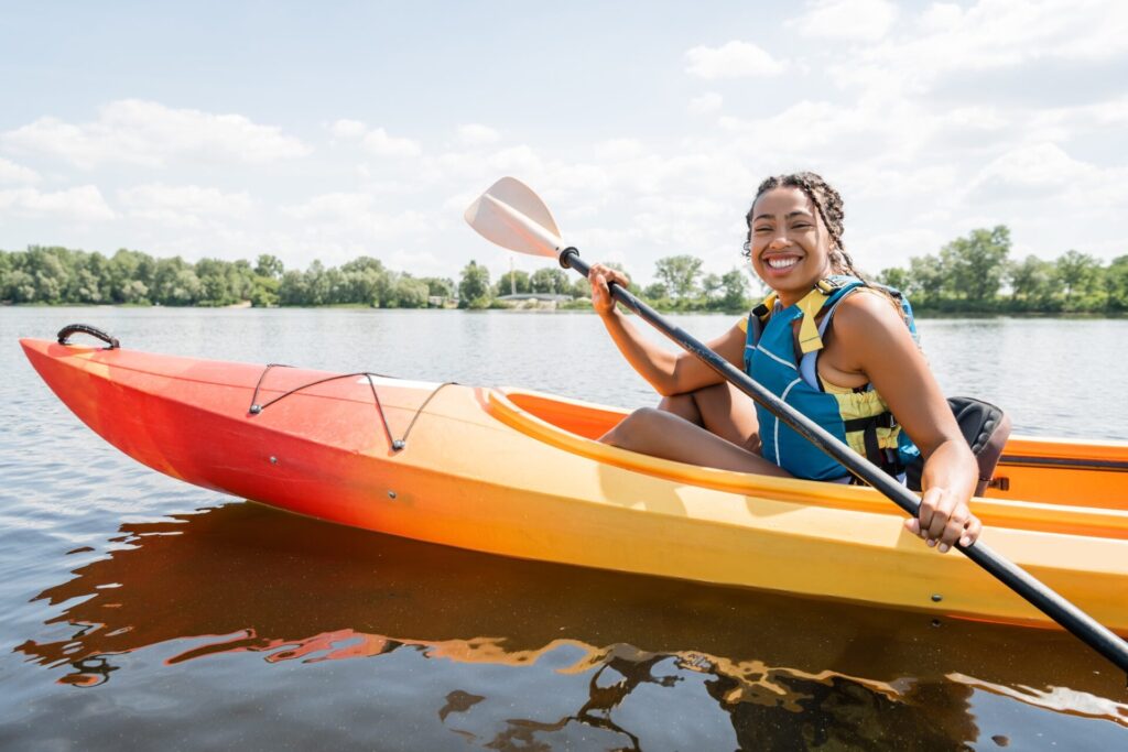 overjoyed african american woman in life vest paddling in sportive kayak and smiling at camera